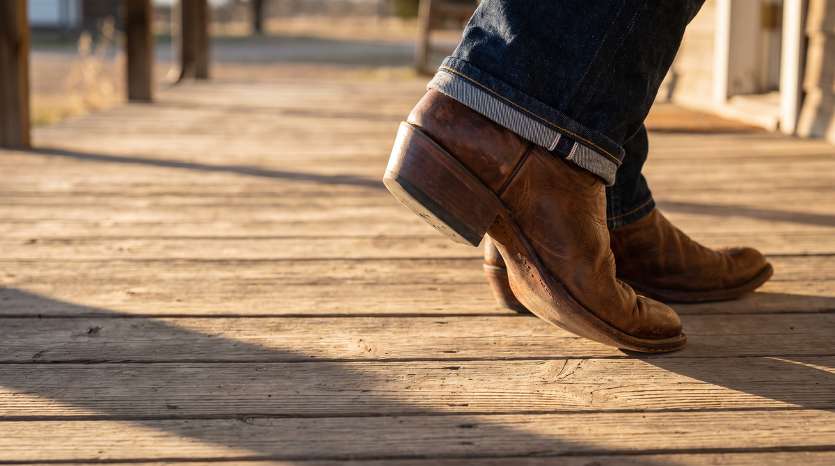 Low angle of a brown cowboy boot mid-step on a wooden porch with the heel lifted slightly off the boards