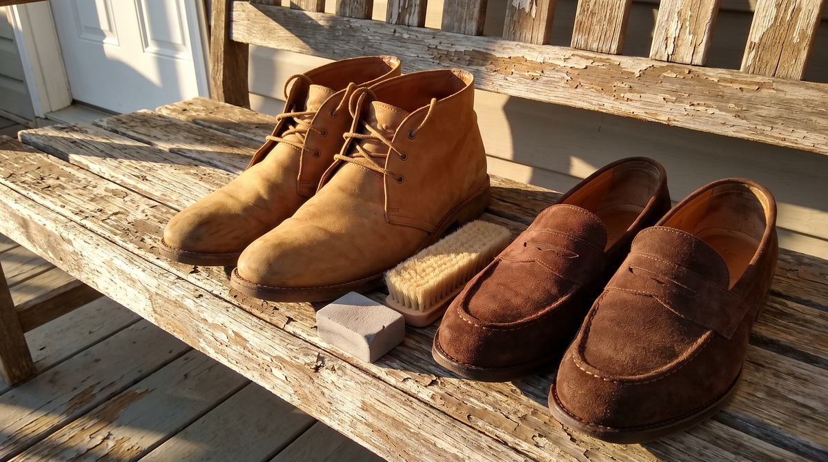 Two tan nubuck chukka boots and a pair of brown suede penny loafers on a porch bench with a brush and an eraser