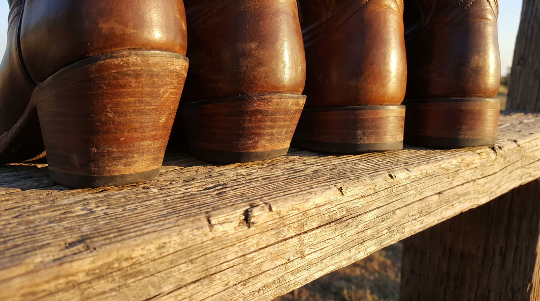 Close-up of the heels of four leather cowboy boots on a wooden plank, from a tall slanted riding heel to lower stacked heels