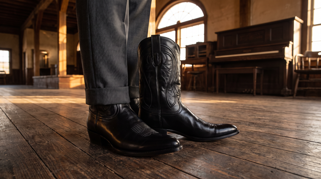 Polished black calfskin cowboy boots under charcoal suit trousers on a dark wood floor in an old Texas dance hall
