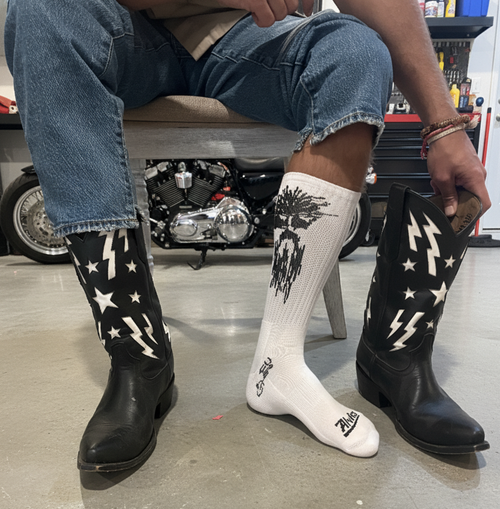 A man wears BFG Boot Socks with cowboy boots beside a motorcycle in a garage.