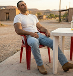 Man in The Shoal Creek tan boots seated outside an adobe building at dusk