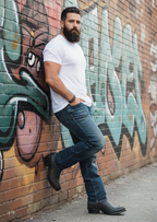 Bearded man in The Shoal Creek boots leaning against a graffiti-covered brick wall