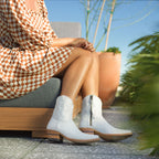 Woman in an orange checkered dress sits outdoors wearing white Trinity ankle boots