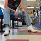Woman sits in a laundromat wearing jeans and white Trinity boots among rows of dryers