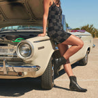 Woman in a checkered mini dress leans on a vintage car wearing black Trinity ankle boots