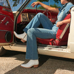 Woman sits in a vintage car's red interior, one leg out the door in white Trinity boots