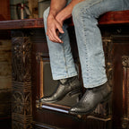 Man seated at a bar, jeans cuffed above The 6th Street black python boots.