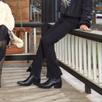 Woman in black jeans and a jacket sits on a porch railing wearing black Trinity boots